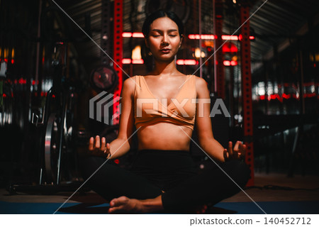 Focused Asian woman meditating cross-legged on a yoga mat in a dark fitness gym, eyes closed for mindful breathing, calm recovery, mental balance, wellness routine, and post workout relaxation. Focused Asian woman meditating cross-legged on a yoga mat in a dark fitness gym, eyes closed for mindful breathing, calm recovery, mental balance, wellness routine, and post workout relaxation. 140452712