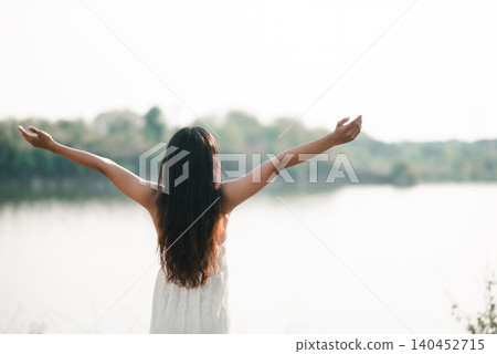 Woman in a white dress standing by a peaceful lake with open arms in soft morning light, showing freedom, nature wellness, fresh air, gratitude, calm lifestyle, travel escape, and emotional release. Woman in a white dress standing by a peaceful lake with open arms in soft morning light, showing freedom, nature wellness, fresh air, gratitude, calm lifestyle, travel escape, and emotional release. 140452715