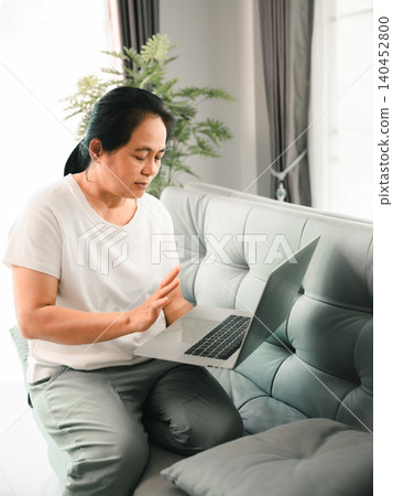Asian woman sitting on a sofa using a laptop at home and raising a hand, showing remote work, video call, online meeting, digital communication, home office, productivity, and modern lifestyle. Asian woman sitting on a sofa using a laptop at home and raising a hand, showing remote work, video call, online meeting, digital communication, home office, productivity, and modern lifestyle. 140452800