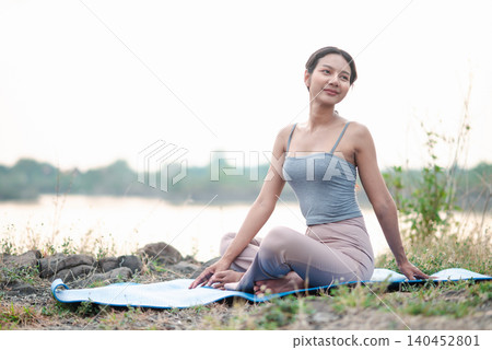 Asian woman sitting on a blue yoga mat beside a quiet lake, resting after practice with a gentle smile, showing outdoor fitness, mindful stretching, relaxation, nature wellness, and peaceful balance. Asian woman sitting on a blue yoga mat beside a quiet lake, resting after practice with a gentle smile, showing outdoor fitness, mindful stretching, relaxation, nature wellness, and peaceful balance. 140452801