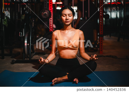 Young athletic Asian woman meditating cross-legged on yoga mat inside a dark gym with eyes closed, showing mindfulness, fitness recovery, balance, breathing, wellness, calm focus, Young athletic Asian woman meditating cross-legged on yoga mat inside a dark gym with eyes closed, showing mindfulness, fitness recovery, balance, breathing, wellness, calm focus, 140452836