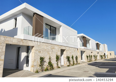 Modern luxury villas with minimalist architecture, natural stone walls, and expansive glass windows in a premium residential community under a bright blue sky. Modern luxury villas with minimalist architecture, natural stone walls, and expansive glass windows in a premium residential community under a bright blue sky. 140453294