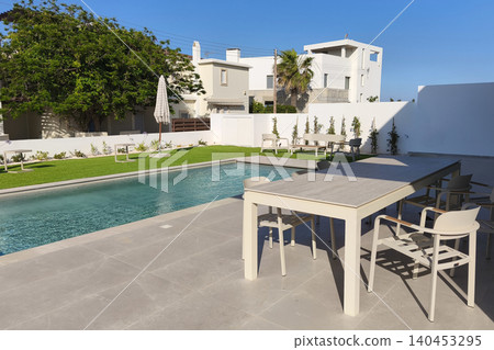 Modern backyard with a swimming pool, patio dining area, and landscaped garden under a clear blue sky, offering a relaxing outdoor lifestyle in a contemporary residential setting. Modern backyard with a swimming pool, patio dining area, and landscaped garden under a clear blue sky, offering a relaxing outdoor lifestyle in a contemporary residential setting. 140453295