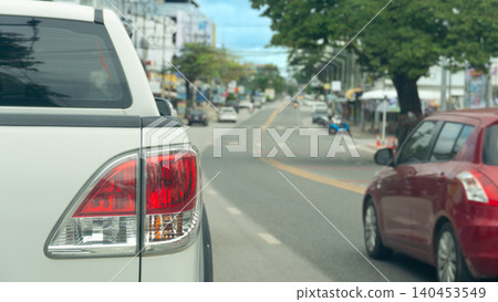 Rear corner close-up of a white truck showing its large tail light driving on asphalt road. A red car travels down the opposing lane of a wide urban road flanked by green trees under a bright sky. Rear corner close-up of a white truck showing its large tail light driving on asphalt road. A red car travels down the opposing lane of a wide urban road flanked by green trees under a bright sky. 140453549