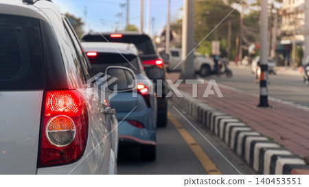 Tight rear view focusing on a white car illuminated red tail light during a traffic jam. A blue car and other commuter vehicles stand queueing on the busy urban roadside lane. Driving on asphalt road. Tight rear view focusing on a white car illuminated red tail light during a traffic jam. A blue car and other commuter vehicles stand queueing on the busy urban roadside lane. Driving on asphalt road. 140453551