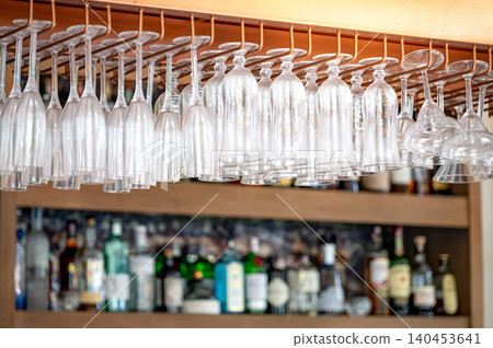 Different empty glasses hang over the bar counter with shelves filled with bottles of alcohol behind Different empty glasses hang over the bar counter with shelves filled with bottles of alcohol behind 140453641