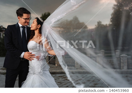 Interracial wedding couple embracing as bride's veil blows 140453960