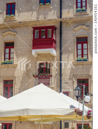 Historic limestone facade with red Maltese gallarija balcony, windows and flower boxes above cafe umbrella in Valletta Malta. High quality photo 140454161