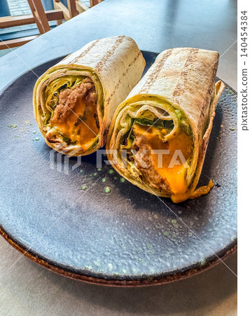 Grilled tortilla wraps cut in half on a black ceramic plate. Two burrito halves filled with meat, lettuce, and melted cheese. Grilled tortilla wraps cut in half on a black ceramic plate. Two burrito halves filled with meat, lettuce, and melted cheese. 140454384