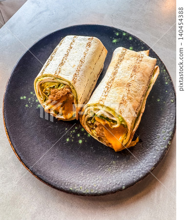 Grilled tortilla wraps cut in half on a black ceramic plate. Two burrito halves filled with meat, lettuce, and melted cheese. Grilled tortilla wraps cut in half on a black ceramic plate. Two burrito halves filled with meat, lettuce, and melted cheese. 140454388