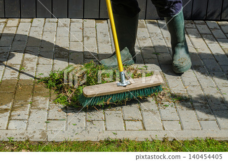 Gardener in Green Rubber Boots Sweeping Conifer Clippings on Stone Paved Walkway with Industrial Turquoise Push Broom Outdoors 140454405
