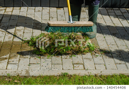 Gardener in Green Rubber Boots Sweeping Conifer Clippings on Stone Paved Walkway with Industrial Turquoise Push Broom Outdoors Gardener in Green Rubber Boots Sweeping Conifer Clippings on Stone Paved Walkway with Industrial Turquoise Push Broom Outdoors 140454406