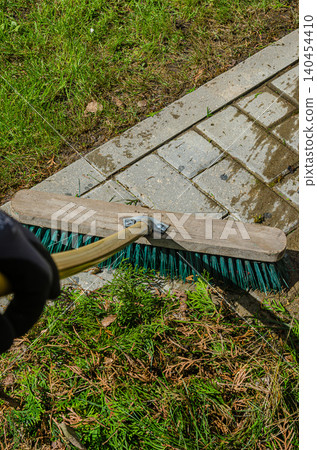 Close-up of a person cleaning a stone walkway. Sweeping green plant waste and branches for a tidy backyard. Close-up of a person cleaning a stone walkway. Sweeping green plant waste and branches for a tidy backyard. 140454410