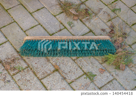 A wooden push broom with green plastic bristles lies flat on outdoor patio pavers amidst scattered dry grass, dead leaves, and yard maintenance debris.. High quality photo A wooden push broom with green plastic bristles lies flat on outdoor patio pavers amidst scattered dry grass, dead leaves, and yard maintenance debris.. High quality photo 140454418
