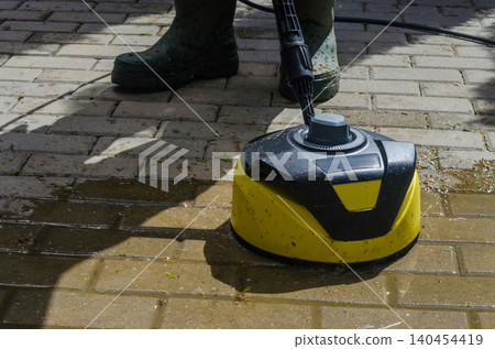 Professional Patio Maintenance: Yellow Pressure Washer Surface Cleaner Scrubbing Wet Stone Tiles with Worker in Rubber Boots 140454419