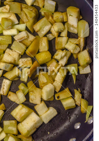 Top-down close up of freshly chopped, pale green vegetable cubes, likely eggplant or zucchini, frying in oil inside a dark non-stick skillet or pan.. High quality photo Top-down close up of freshly chopped, pale green vegetable cubes, likely eggplant or zucchini, frying in oil inside a dark non-stick skillet or pan.. High quality photo 140454433