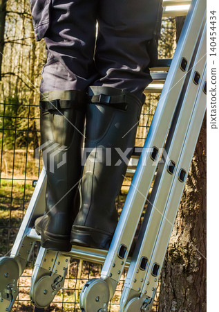 Person in rubber boots stands on an aluminum ladder for outdoor garden maintenance and home improvement projects in a sunny yard setting. High quality photo Person in rubber boots stands on an aluminum ladder for outdoor garden maintenance and home improvement projects in a sunny yard setting. High quality photo 140454434
