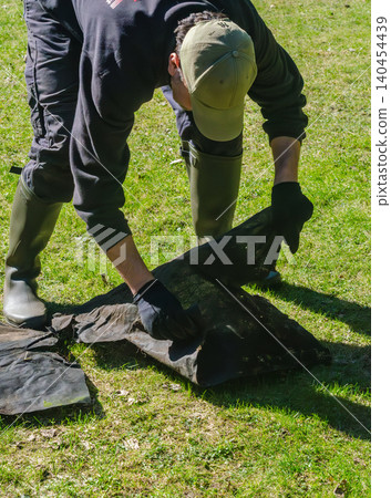 Gardener in boots laying black landscape fabric weed barrier on green lawn for backyard garden renovation and maintenance. Gardener in boots laying black landscape fabric weed barrier on green lawn for backyard garden renovation and maintenance. 140454439