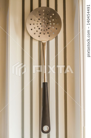 A well-used metal skimmer spoon, a common kitchen tool for straining or frying, hangs against a modern, clean, white grooved wall. High quality photo 140454491