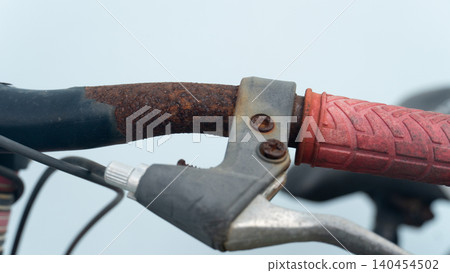 Detailed shot of an old bicycle's handlebar featuring a rusty metal stem, a silver brake lever mechanism, and a textured red rubber grip handle set against a blurry backdrop. Detailed shot of an old bicycle's handlebar featuring a rusty metal stem, a silver brake lever mechanism, and a textured red rubber grip handle set against a blurry backdrop. 140454502