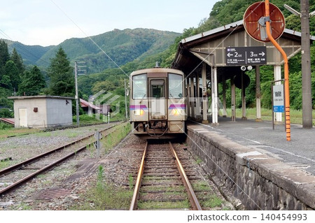 一列 Kiha 120 系列本地列車行駛在花備線上，停靠在備後落合站的月台上，周圍綠樹環繞。 140454993