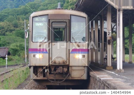 一列 Kiha 120 系列本地列車行駛在花備線上，停靠在備後落合站的月台上，周圍綠樹環繞。 140454994