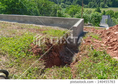 Groundwork and drainage trenching on a residential construction site. Long narrow pit dug next to concrete retaining wall under clear day light with green nature background Groundwork and drainage trenching on a residential construction site. Long narrow pit dug next to concrete retaining wall under clear day light with green nature background 140455763
