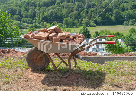Horizontal panoramic shot of a metal construction wheelbarrow filled with broken red bricks on a hillside. Wide green valley, forest, and blue hills form the summer backdrop Horizontal panoramic shot of a metal construction wheelbarrow filled with broken red bricks on a hillside. Wide green valley, forest, and blue hills form the summer backdrop 140455769