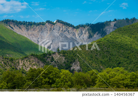 Wide natural landscape with steep rocky cliffs, lush green hills, dense trees in the foreground, and bright daylight under a clear blue sky Wide natural landscape with steep rocky cliffs, lush green hills, dense trees in the foreground, and bright daylight under a clear blue sky 140456367