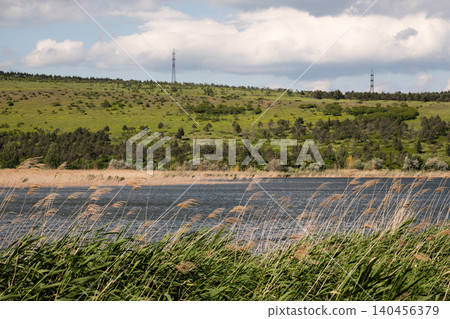 A wide summer landscape featuring a blue lake, lush green reeds, and rolling hills under a cloudy sky A wide summer landscape featuring a blue lake, lush green reeds, and rolling hills under a cloudy sky 140456379