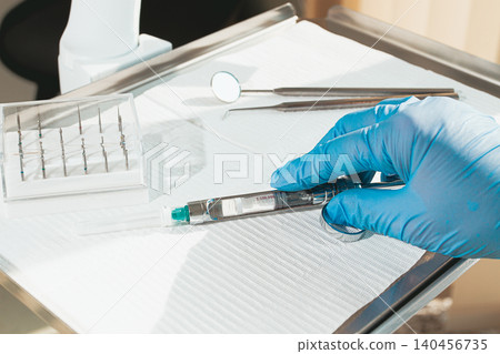 The dentist's hand, in a blue glove, holds a carpal syringe on a tray, ready for a medical procedure. The dentist's hand, in a blue glove, holds a carpal syringe on a tray, ready for a medical procedure. 140456735