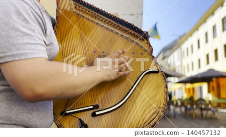 Male fingers of musician playing the bandura on a sunny summer day. Sunlight falls on the strings and wood surface, creating a warm and lively scene of outdoor music and authentic street performance Male fingers of musician playing the bandura on a sunny summer day. Sunlight falls on the strings and wood surface, creating a warm and lively scene of outdoor music and authentic street performance 140457132