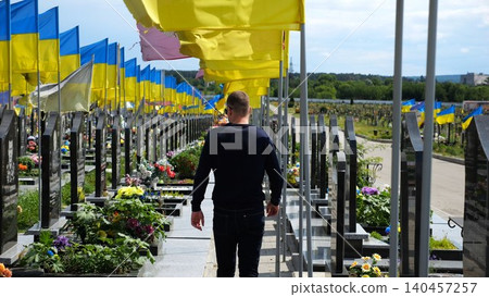 Mourning man go among countless graves of ukrainian soldiers with national flags at military cemetery in Kharkiv. Scene reflects personal sorrow and nation collective loss caused by russian aggression Mourning man go among countless graves of ukrainian soldiers with national flags at military cemetery in Kharkiv. Scene reflects personal sorrow and nation collective loss caused by russian aggression 140457257