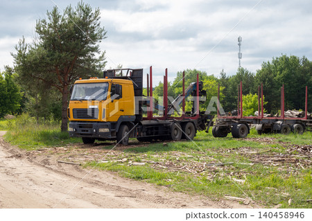 Timber truck with crane trailer at logging industry yard, yellow logging vehicle parked near forest on cloudy day Timber truck with crane trailer at logging industry yard, yellow logging vehicle parked near forest on cloudy day 140458946