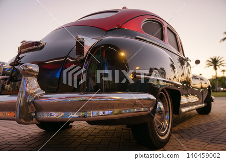Low angle rear view of a black and red vintage taxi style automobile with chrome bumper and retro body design. Historic classic transportation vehicle is parked outdoors during sunset with palm trees Low angle rear view of a black and red vintage taxi style automobile with chrome bumper and retro body design. Historic classic transportation vehicle is parked outdoors during sunset with palm trees 140459002