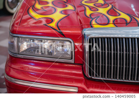Close-up view of a striking red car featuring custom flame graphics in yellow and red. The chrome grille and headlight assembly enhance the vehicle's bold aesthetic, showcasing its unique design. Close-up view of a striking red car featuring custom flame graphics in yellow and red. The chrome grille and headlight assembly enhance the vehicle's bold aesthetic, showcasing its unique design. 140459005
