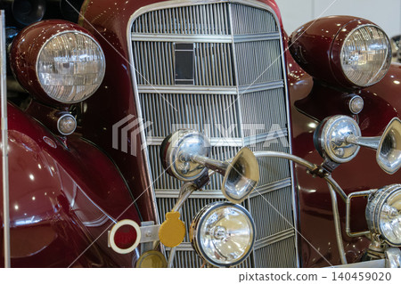 Close-up view captures front of vintage dark red automobile. Focus on classic grille with vertical slats, dual round headlights, and vintage chrome horns, showcasing intricate automotive design. Close-up view captures front of vintage dark red automobile. Focus on classic grille with vertical slats, dual round headlights, and vintage chrome horns, showcasing intricate automotive design. 140459020