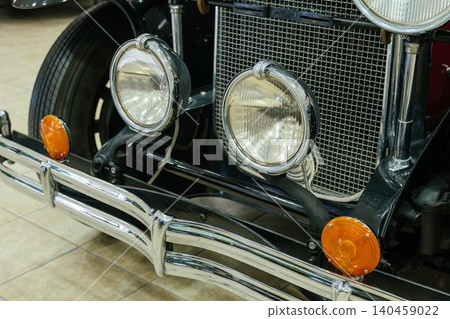 Detailed view of front section of vintage car, highlighting chrome bumper and twin headlights. Bright orange turn signals and metal mesh grille add to classic automotive design. Detailed view of front section of vintage car, highlighting chrome bumper and twin headlights. Bright orange turn signals and metal mesh grille add to classic automotive design. 140459022