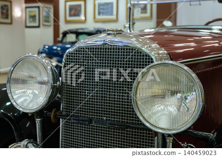 Detailed view captures front of classic maroon automobile showcasing round headlights and intricate chrome grille. Soft background features a blue vintage car and framed artwork, enhancing the elegant Detailed view captures front of classic maroon automobile showcasing round headlights and intricate chrome grille. Soft background features a blue vintage car and framed artwork, enhancing the elegant 140459023