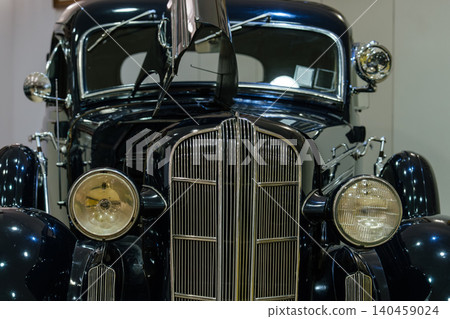 Detailed front view of classic dark blue automobile showcasing chrome grille, circular headlights, and partially open hood. Indoor setting highlights the car's glossy finish and elegant design. Detailed front view of classic dark blue automobile showcasing chrome grille, circular headlights, and partially open hood. Indoor setting highlights the car's glossy finish and elegant design. 140459024