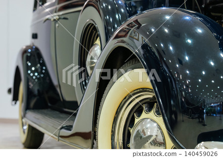 Detailed close-up captures shiny fender and white-walled tire of vintage dark blue car. Polished surface reflects lights and framed artwork, enhancing visual appeal in an exhibition setting. Detailed close-up captures shiny fender and white-walled tire of vintage dark blue car. Polished surface reflects lights and framed artwork, enhancing visual appeal in an exhibition setting. 140459026
