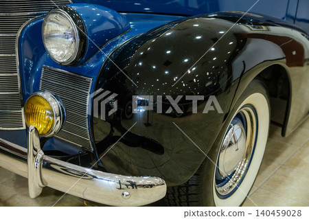 Detailed low-angle view captures front fender and headlight of classic blue automobile. Glossy black fender showcases reflections of interior lights and other vehicles, suggesting museum setting. 140459028