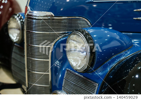 Detailed close-up showcases front right side of vintage blue automobile. Focus on round headlight with chrome rim and multi-slat chrome grille, reflecting studio lights. Detailed close-up showcases front right side of vintage blue automobile. Focus on round headlight with chrome rim and multi-slat chrome grille, reflecting studio lights. 140459029
