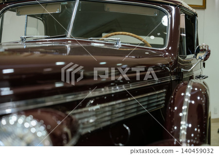 Close-up shot captures vintage maroon automobile showcasing shiny paint and chrome details. Interior features wooden steering wheel and light-colored fabric, suggesting luxury in a museum setting. 140459032