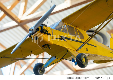 Close-up view of a vintage yellow small airplane hanging from the ceiling indoors. Features include a silver propeller, transparent cockpit, and black markings, with sunlight illuminating the scene. Close-up view of a vintage yellow small airplane hanging from the ceiling indoors. Features include a silver propeller, transparent cockpit, and black markings, with sunlight illuminating the scene. 140459044