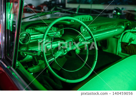 Close-up view of vintage car's dashboard and steering wheel illuminated by vibrant green light. Retro design features chrome accents, dials, and controls, creating a unique atmosphere. 140459056