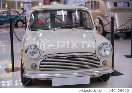 Front view of cream classic compact car showcased in well-lit indoor exhibition space. Prominent chrome grille, circular headlights, and polished floor reflect vintage vehicles and display barriers. Front view of cream classic compact car showcased in well-lit indoor exhibition space. Prominent chrome grille, circular headlights, and polished floor reflect vintage vehicles and display barriers. 140459060