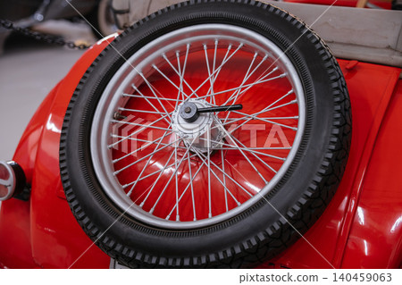 Detailed close-up of rear-mounted spare tire on vintage red automobile. Tire features classic tread design, mounted on silver wire-spoke wheel, showcasing vibrant paint and intricate details. Detailed close-up of rear-mounted spare tire on vintage red automobile. Tire features classic tread design, mounted on silver wire-spoke wheel, showcasing vibrant paint and intricate details. 140459063
