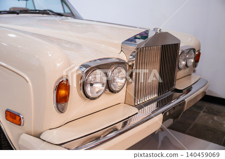 Frontal close-up of antique cream-colored luxury car showcasing iconic chrome grille and dual round headlights. Bright lighting highlights polished surfaces against a clean background. Frontal close-up of antique cream-colored luxury car showcasing iconic chrome grille and dual round headlights. Bright lighting highlights polished surfaces against a clean background. 140459069