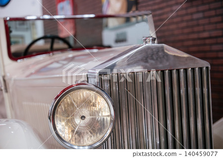 Close-up, low-angle shot highlights front grille and headlight of vintage cream car. Polished chrome accents and vertical ribs suggest classic automotive design, with a soft-focus brick wall in Close-up, low-angle shot highlights front grille and headlight of vintage cream car. Polished chrome accents and vertical ribs suggest classic automotive design, with a soft-focus brick wall in 140459077